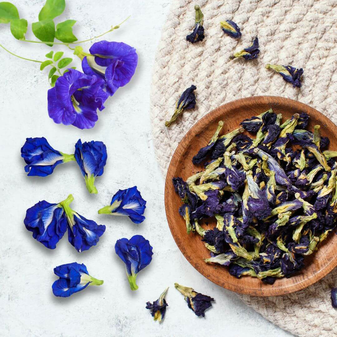Dried butterfly pea flowers in a wooden bowl with flowers scattered beside it